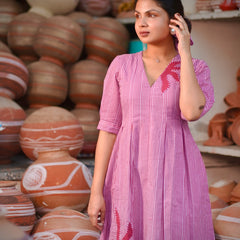 Woman wearing a pink 100% cotton gingham maxi dress with artistic sun motif print, V-neck, three-quarter sleeves, and side pockets, standing among handcrafted clay pottery.