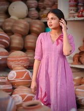 Woman wearing a pink 100% cotton gingham maxi dress with artistic sun motif print, V-neck, three-quarter sleeves, and side pockets, standing among handcrafted clay pottery.