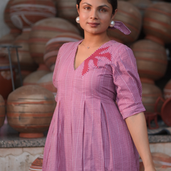 Woman wearing a pink 100% cotton gingham maxi dress with artistic sun motif print, V-neck, three-quarter sleeves, and side pockets, standing among handcrafted clay pottery.