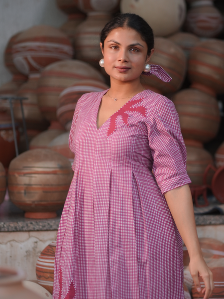 Woman wearing a pink 100% cotton gingham maxi dress with artistic sun motif print, V-neck, three-quarter sleeves, and side pockets, standing among handcrafted clay pottery.