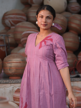 Woman wearing a pink 100% cotton gingham maxi dress with artistic sun motif print, V-neck, three-quarter sleeves, and side pockets, standing among handcrafted clay pottery.