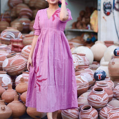 Woman wearing a pink 100% cotton gingham maxi dress with artistic sun motif print, V-neck, three-quarter sleeves, and side pockets, standing among handcrafted clay pottery.