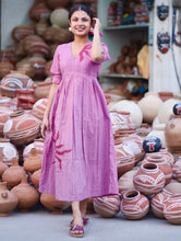 Woman wearing a pink 100% cotton gingham maxi dress with artistic sun motif print, V-neck, three-quarter sleeves, and side pockets, standing among handcrafted clay pottery.