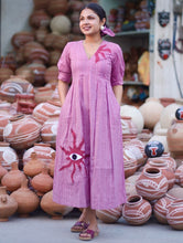 Woman wearing a pink 100% cotton gingham maxi dress with artistic sun motif print, V-neck, three-quarter sleeves, and side pockets, standing among handcrafted clay pottery.