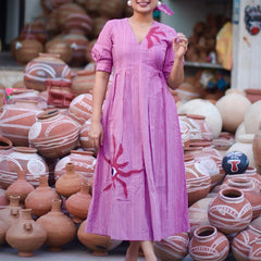 Woman wearing a pink 100% cotton gingham maxi dress with artistic sun motif print, V-neck, three-quarter sleeves, and side pockets, standing among handcrafted clay pottery.
