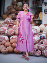 Woman wearing a pink 100% cotton gingham maxi dress with artistic sun motif print, V-neck, three-quarter sleeves, and side pockets, standing among handcrafted clay pottery.