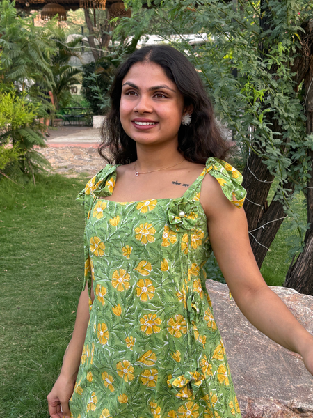 Woman wearing a green floral A-line dress with a square neckline, adjustable tie-up shoulder straps, and knee-length silhouette in a garden setting.