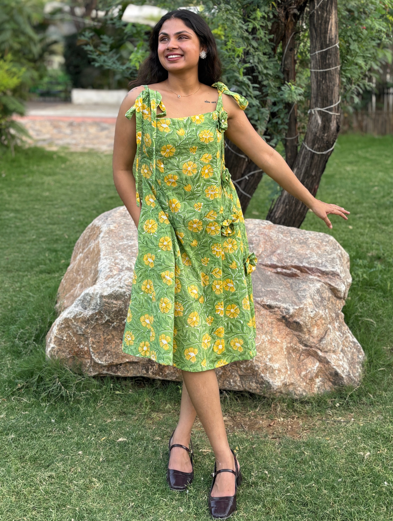 Woman wearing a green floral A-line dress with a square neckline, adjustable tie-up shoulder straps, and knee-length silhouette in a garden setting.