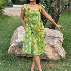 Woman wearing a green floral A-line dress with a square neckline, adjustable tie-up shoulder straps, and knee-length silhouette in a garden setting.
