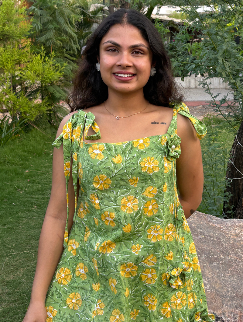 Woman wearing a green floral A-line dress with a square neckline, adjustable tie-up shoulder straps, and knee-length silhouette in a garden setting.