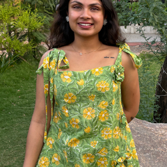 Woman wearing a green floral A-line dress with a square neckline, adjustable tie-up shoulder straps, and knee-length silhouette in a garden setting.